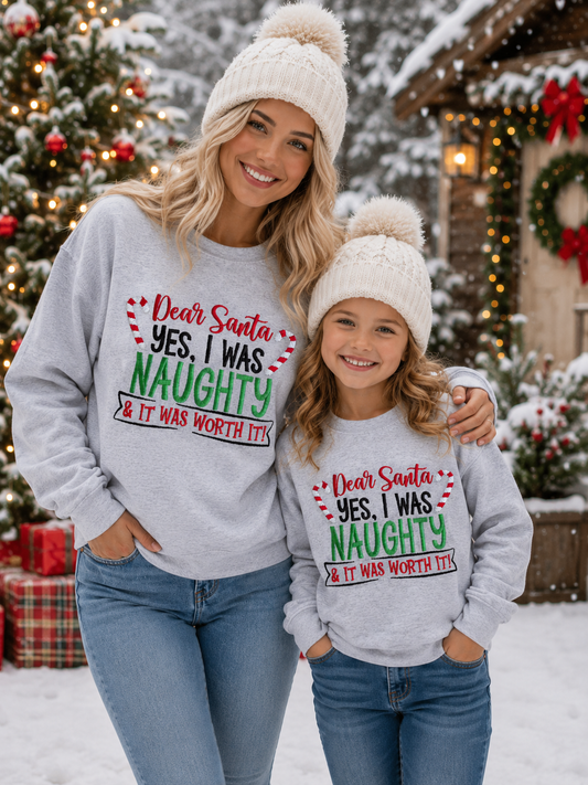 A woman and a young girl wearing matching light gray Christmas sweatshirts and white knit pom-pom beanies, standing outdoors in the snow in front of decorated Christmas trees and a house. The sweatshirts have a festive graphic that reads 'Dear Santa, YES, I WAS NAUGHTY & IT WAS WORTH IT!' in red, green, and black text.