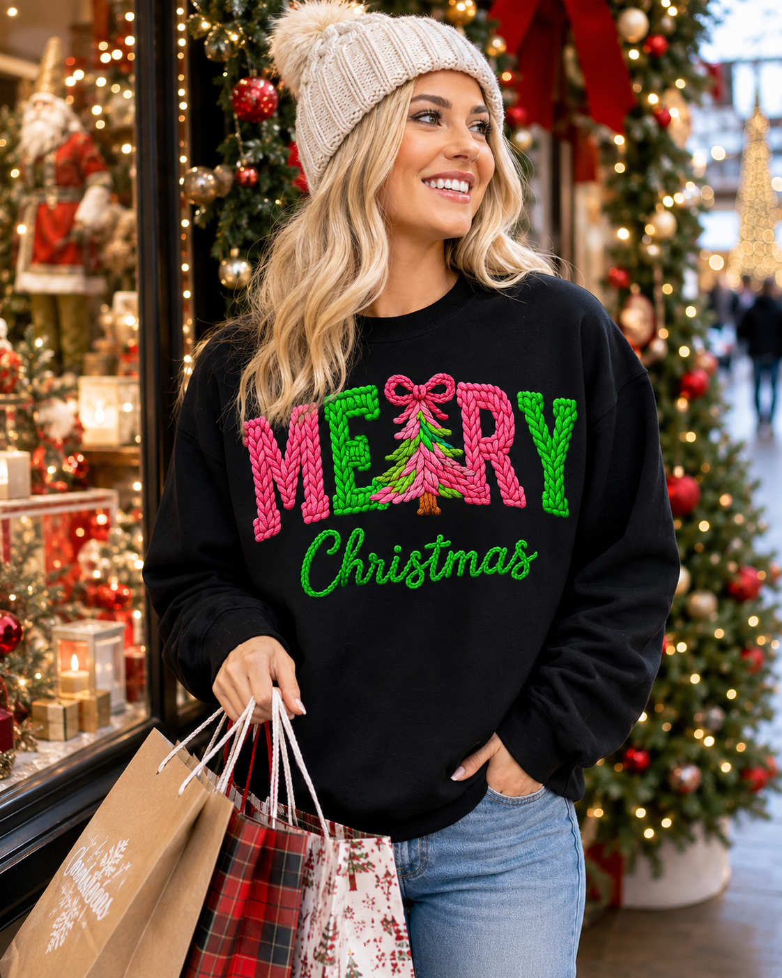 A woman wearing a black crewneck sweatshirt with a 'MERRY Christmas' graphic in pink and green, styled with a beige knit beanie and blue jeans, holding shopping bags in front of a decorated Christmas tree store window.
