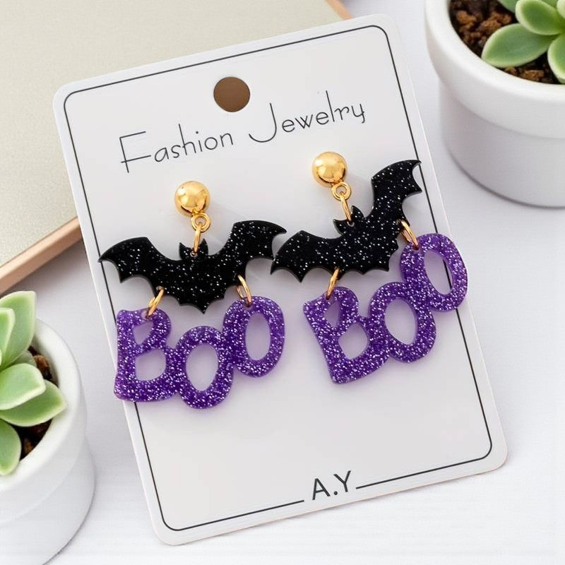 Halloween-themed earrings with bats and 'BOO' on a card, placed on a white surface with plants.