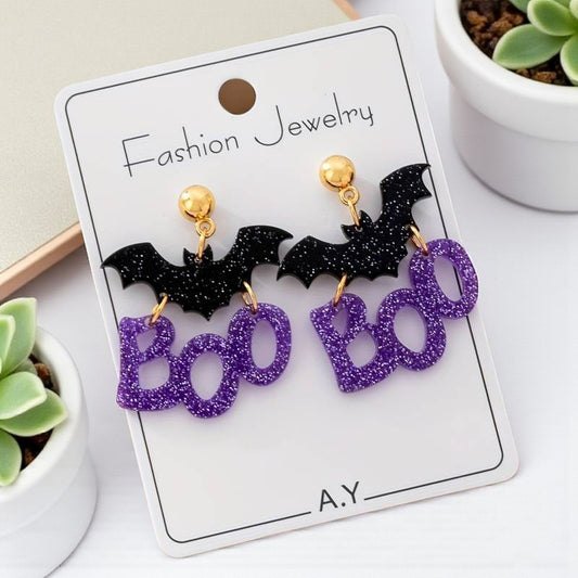 Halloween-themed earrings with bats and 'BOO' on a card, placed on a white surface with plants.